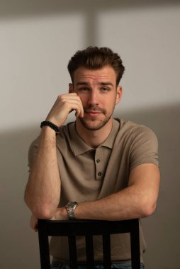 Tecrit edilmiş arka planda oturan yakışıklı bir gencin stüdyo fotoğrafı. Polo tişört giyen ve kameraya bakan kendinden emin bir erkek. Uzayı kopyala İzole edilmiş arkaplan. 