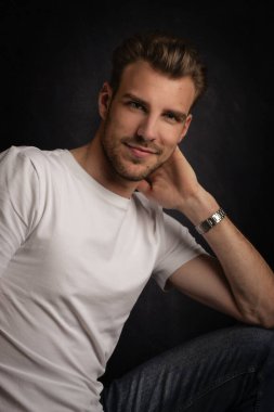 İzole edilmiş karanlık arka planda oturan genç bir adamın stüdyo görüntüsü. Beyaz tişört giyen kendinden emin bir erkek. Boşluğu kopyala.