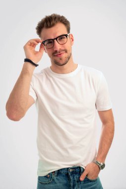 İzole edilmiş arka planda duran genç bir adamın stüdyo çekimi. Beyaz tişört giyen ve okuma gözlüğü takan kendinden emin bir erkek. Uzayı kopyala Elleriyle bardaklara dokunuyor.. 