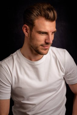 İzole edilmiş karanlık arka planda oturan genç bir adamın stüdyo görüntüsü. Beyaz tişört giyen kendinden emin bir erkek. Boşluğu kopyala.