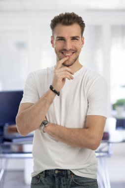 Modern bir ofiste tek başına dururken gülümseyen kendinden emin genç bir iş adamının portresi. Gündelik kıyafetler giyen genç bir girişimci. Eller çenede.. 
