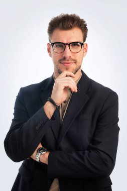 Beyaz arka planda duran yakışıklı bir iş adamının stüdyo fotoğrafı. Kendine güvenen erkek polo gömlek ve ceket giyiyor ve düşünceli görünüyor. Uzayı kopyala İzole edilmiş arka plan. Eller çenede..