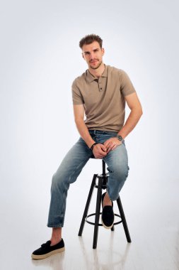 Gri arka planda oturan yakışıklı bir gencin stüdyo fotoğrafı. Polo tişört giyen ve kameraya gülümseyen kendinden emin bir erkek. Uzayı kopyala İzole edilmiş arka plan. Tam uzunluk. 