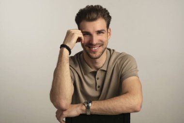 Gri arka planda oturan yakışıklı bir gencin stüdyo fotoğrafı. Polo tişört giyen ve kameraya gülümseyen kendinden emin bir erkek. Uzayı kopyala İzole edilmiş arkaplan. 