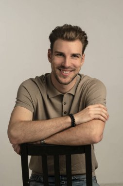 Gri arka planda oturan yakışıklı bir gencin stüdyo fotoğrafı. Polo tişört giyen ve kameraya gülümseyen kendinden emin bir erkek. Uzayı kopyala İzole edilmiş arkaplan. 