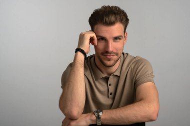 Gri arka planda oturan yakışıklı bir gencin stüdyo fotoğrafı. Polo tişört giyen ve kameraya gülümseyen kendinden emin bir erkek. Uzayı kopyala İzole edilmiş arkaplan. 