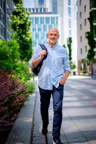 Şehir caddesinde yürüyen orta yaşlı bir adam. Tişört ve sırt çantası giyen kendinden emin bir erkek. Tam boy çekim.