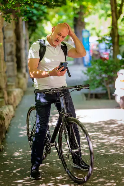 Şehirde bisikletli bir banliyö. Son sınıf öğrencisi sokakta bisiklet sürüyor ve akıllı telefonunu kullanıyor. Gündelik giysiler ve sırt çantası giyen kendinden emin bir erkek. Tam boy çekim.