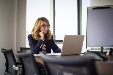 Sarı saçlı iş kadını dizüstü bilgisayar kullanıyor ve iş yerinde internet konferansı yapıyor. Olgun bir bayan patronun portresi, müdür ve CEO internette geziniyor ve online planlama yapıyor. İş kıyafeti giyen çekici bir kadın.. 