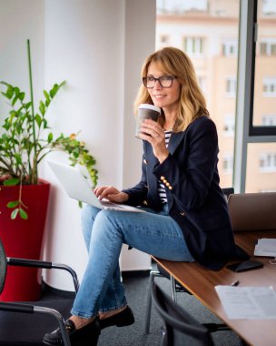 Laptop kullanan, iş yerinde kahve içen sarı saçlı bir iş kadını. Olgun bir bayan patronun portresi, müdür ve CEO internette geziniyor ve online planlama yapıyor. İş kıyafeti giyen çekici bir kadın.. 