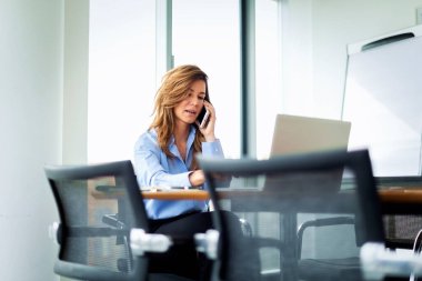 Sarı saçlı iş kadını dizüstü bilgisayarın önünde oturuyor ve telefon ediyor. Kendine güvenen bir kadın iş kıyafeti giyiyor ve modern bir ofiste çalışıyor.. 