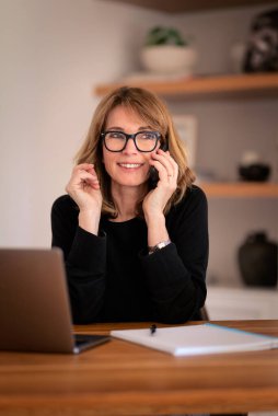 Evde dizüstü bilgisayarı ve akıllı telefonu başında oturan sarı saçlı bir kadın. Evden çalışan orta yaşlı beyaz bir kadın. Merkez Ofis. 