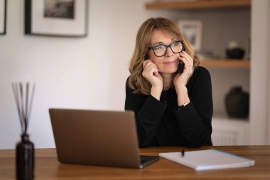 Evde dizüstü bilgisayarı ve akıllı telefonu başında oturan sarı saçlı bir kadın. Evden çalışan orta yaşlı beyaz bir kadın. Merkez Ofis. 