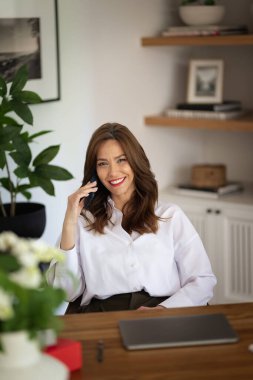 Kumral saçlı bir kadın evde dizüstü bilgisayarının önünde oturuyor ve telefonda konuşuyor. Beyaz gömlek giyen ve evden çalışan orta yaşlı bir kadın. Merkez Ofis. 