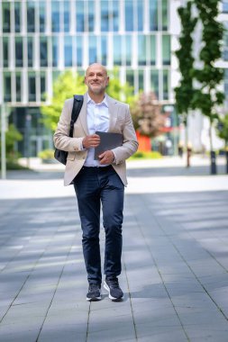 Diz üstü bilgisayarı ve sırt çantası olan orta yaşlı bir adam şehir caddesinde yürüyor. Kendine güveni tam bir iş adamı. Tam boy çekim. 