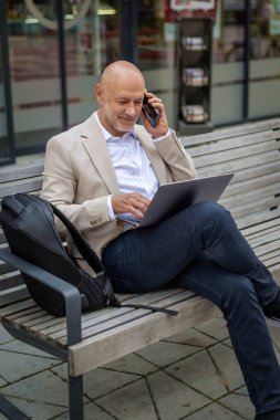 Şehirdeki bir bankta oturan yakışıklı iş adamı cep telefonu ve dizüstü bilgisayar kullanıyor. Profesyonel erkek iş kıyafeti giyiyor ve çalışıyor.. 