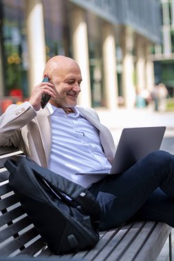 Şehirdeki bir bankta oturan yakışıklı iş adamı cep telefonu ve dizüstü bilgisayar kullanıyor. Profesyonel erkek iş kıyafeti giyiyor ve çalışıyor.. 