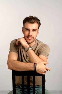 İzole edilmiş arka planda bir sandalyede oturan genç bir adamın stüdyo fotoğrafı. Tişört giyen ve kameraya bakan kendinden emin bir erkek. Boşluğu kopyala.