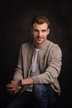 İzole edilmiş siyah arka planda oturan genç bir adamın stüdyo fotoğrafı. Kendine güvenen, ceketli ve kameraya bakan bir erkek. Boşluğu kopyala.