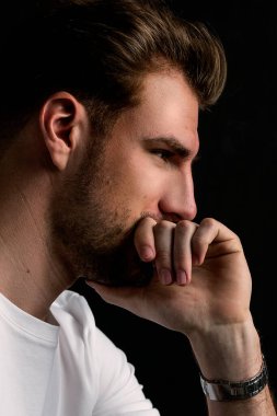 İzole edilmiş karanlık arka planda oturan genç bir adamın stüdyo görüntüsü. Beyaz tişört giyen kendinden emin bir erkek. Boşluğu kopyala.