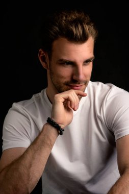 İzole edilmiş karanlık arka planda oturan genç bir adamın stüdyo görüntüsü. Beyaz tişört giyen kendinden emin bir erkek. Boşluğu kopyala.