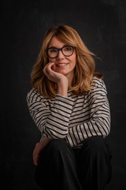 50 'li yaşlarının sonunda gülümseyen sarı saçlı bir kadının portresi. Okuma gözlüğü ve çizgili kazak giyen çekici bir kadın. İzole edilmiş arkaplan. 