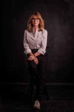 Orta yaşlı bir kadının esmer arka planda tam boy portresi. İzole edilmiş arka planda oturan sarı saçlı kadının stüdyo fotoğrafı. Boşluğu kopyala. 