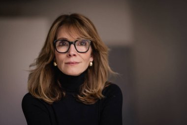 50 'li yaşlarının sonunda gülümseyen sarı saçlı bir kadının portresi. Okuma gözlüğü ve balıkçı yaka kazak giyen çekici bir kadın.. 