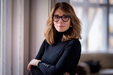 50 'li yaşlarının sonunda gülümseyen sarı saçlı bir kadının portresi. Okuma gözlüğü ve balıkçı yaka kazak giyen çekici bir kadın.. 