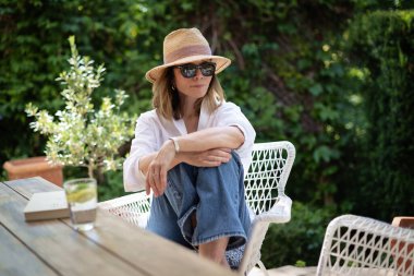 Sarı saçlı kadın arka bahçede oturuyor ve dinleniyor. Beyaz gömlek ve güneş şapkası giyen çekici olgun bir kadın.. 