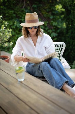 Sarışın kadın arka bahçede oturuyor ve kitap okuyor. Beyaz gömlek ve güneş şapkası giyen çekici olgun bir kadın. Evde dinlenen bir kadın. Tam boy çekim. 