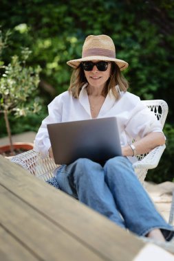 Arka bahçede masada otururken dizüstü bilgisayar kullanan sarı saçlı kadın. Evde çalışan çekici olgun bir kadın. Merkez Ofis. 