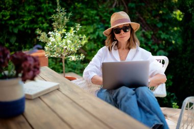 Arka bahçede masada otururken dizüstü bilgisayar kullanan sarı saçlı kadın. Evde çalışan çekici olgun bir kadın. Merkez Ofis. 