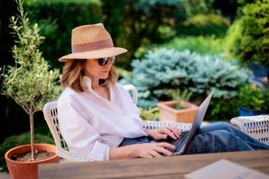 Arka bahçede masada otururken dizüstü bilgisayar kullanan sarı saçlı kadın. Evde çalışan çekici olgun bir kadın. Merkez Ofis. 