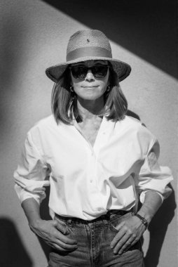 Duvarın önünde poz veren gülümseyen bir kadın resmi. Hasır şapkalı ve güneşli bir kadın. Beyaz gömlek giyen ve kameraya gülümseyen sarı saçlı kadın. Boşluğu kopyala. 