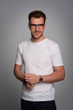 Tecrit edilmiş arka planda duran yakışıklı bir gencin stüdyo fotoğrafı. Beyaz tişört giyen ve kameraya bakan kendinden emin bir erkek. Uzayı kopyala İzole edilmiş arka plan. Eller çenede.. 