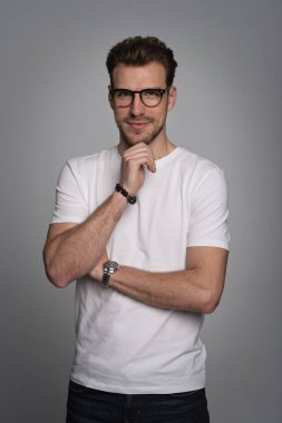 Tecrit edilmiş arka planda duran yakışıklı bir gencin stüdyo fotoğrafı. Beyaz tişört giyen ve kameraya bakan kendinden emin bir erkek. Uzayı kopyala İzole edilmiş arka plan. Eller çenede.. 
