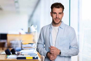 Modern bir ofiste tek başına dururken gülümseyen kendinden emin genç bir iş adamının portresi. Gündelik kıyafetler giyen genç girişimci. 