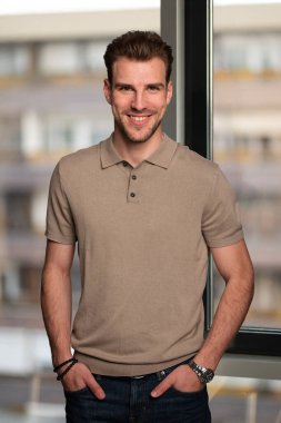 Cam duvarın önünde tek başına dururken gülümseyen kendinden emin bir gencin portresi. Genç erkek, günlük kıyafetler giyiyor.. 