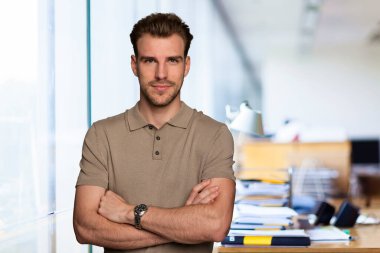 Modern bir ofiste tek başına dururken gülümseyen kendinden emin genç bir iş adamının portresi. Gündelik kıyafetler giyen genç girişimci. 