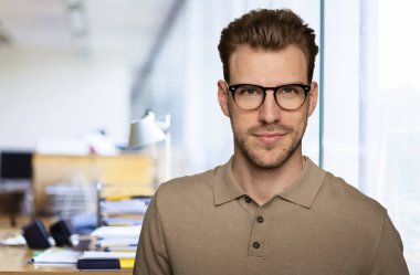 Modern bir ofiste tek başına dururken gülümseyen kendinden emin genç bir iş adamının portresi. Gündelik kıyafetler giyen genç girişimci. 