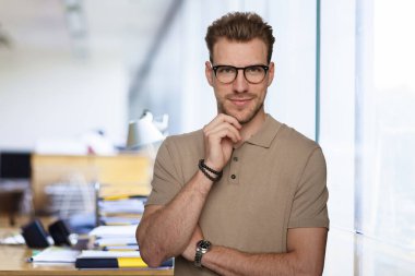 Modern bir ofiste tek başına dururken gülümseyen kendinden emin genç bir iş adamının portresi. Gündelik kıyafetler giyen genç girişimci. 