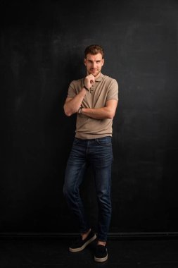 Tecrit edilmiş arka planda duran yakışıklı bir gencin stüdyo fotoğrafı. Polo tişört giyen ve kameraya bakan kendinden emin bir erkek. Uzayı kopyala İzole edilmiş koyu bir arka plan. Eller çenede. Tam uzunluk.