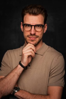 Tecrit edilmiş arka planda duran yakışıklı bir gencin stüdyo fotoğrafı. Polo tişört giyen ve kameraya bakan kendinden emin bir erkek. Uzayı kopyala İzole edilmiş koyu bir arka plan. Eller çenede.. 