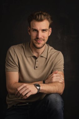 Tecrit edilmiş arka planda oturan yakışıklı bir gencin stüdyo fotoğrafı. Polo tişört giyen ve kameraya bakan kendinden emin bir erkek. Uzayı kopyala İzole edilmiş koyu bir arka plan. Eller çenede. Tam uzunluk.