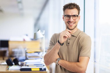 Modern bir ofiste tek başına dururken gülümseyen kendinden emin genç bir iş adamının portresi. Gündelik kıyafetler giyen genç girişimci. 