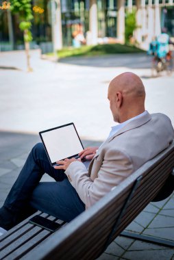 Orta yaşlı bir adam sokakta bir bankta bilgisayar laptopu kullanarak oturuyor. Gündelik kıyafetler giyen kendinden emin bir erkek. Boş ekran. Arka görünüm. 