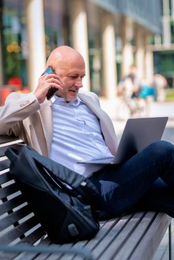 Sokakta bir bankta oturan orta yaşlı bir adam telefon kullanıyor. Gündelik kıyafetler giyen kendinden emin bir erkek.. 