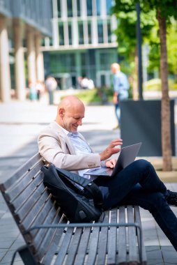 Orta yaşlı bir adam sokakta bir bankta bilgisayar laptopu kullanarak oturuyor. Gündelik kıyafetler giyen kendinden emin bir erkek.. 