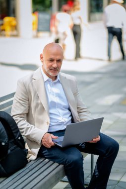 Orta yaşlı bir adam sokakta bir bankta bilgisayar laptopu kullanarak oturuyor. Gündelik kıyafetler giyen kendinden emin bir erkek.. 